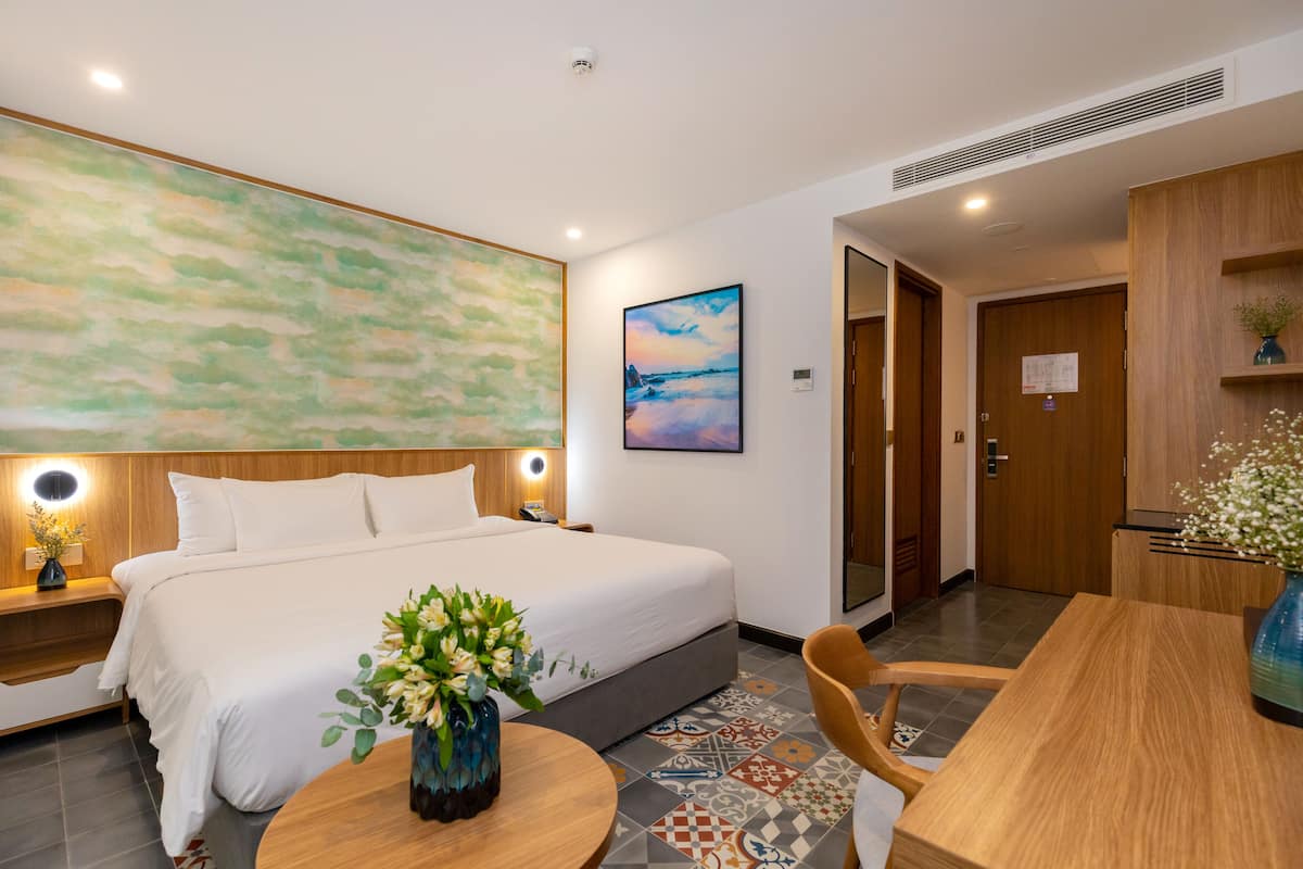 A hotel room with a large bed, a round wooden table, a vase of flowers, and a painting on the wall.