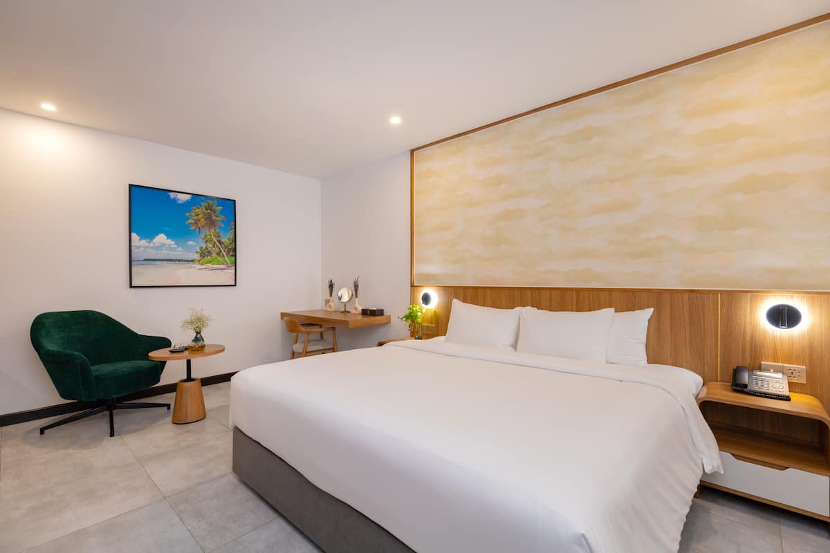 A modern hotel room with a large bed, a desk, a green armchair, and a painting of a beach scene.