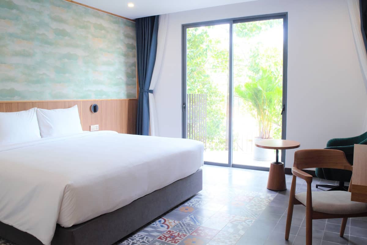 A hotel room with a large bed, a wooden headboard, a sliding glass door to the balcony, a small round table, and a wooden chair.