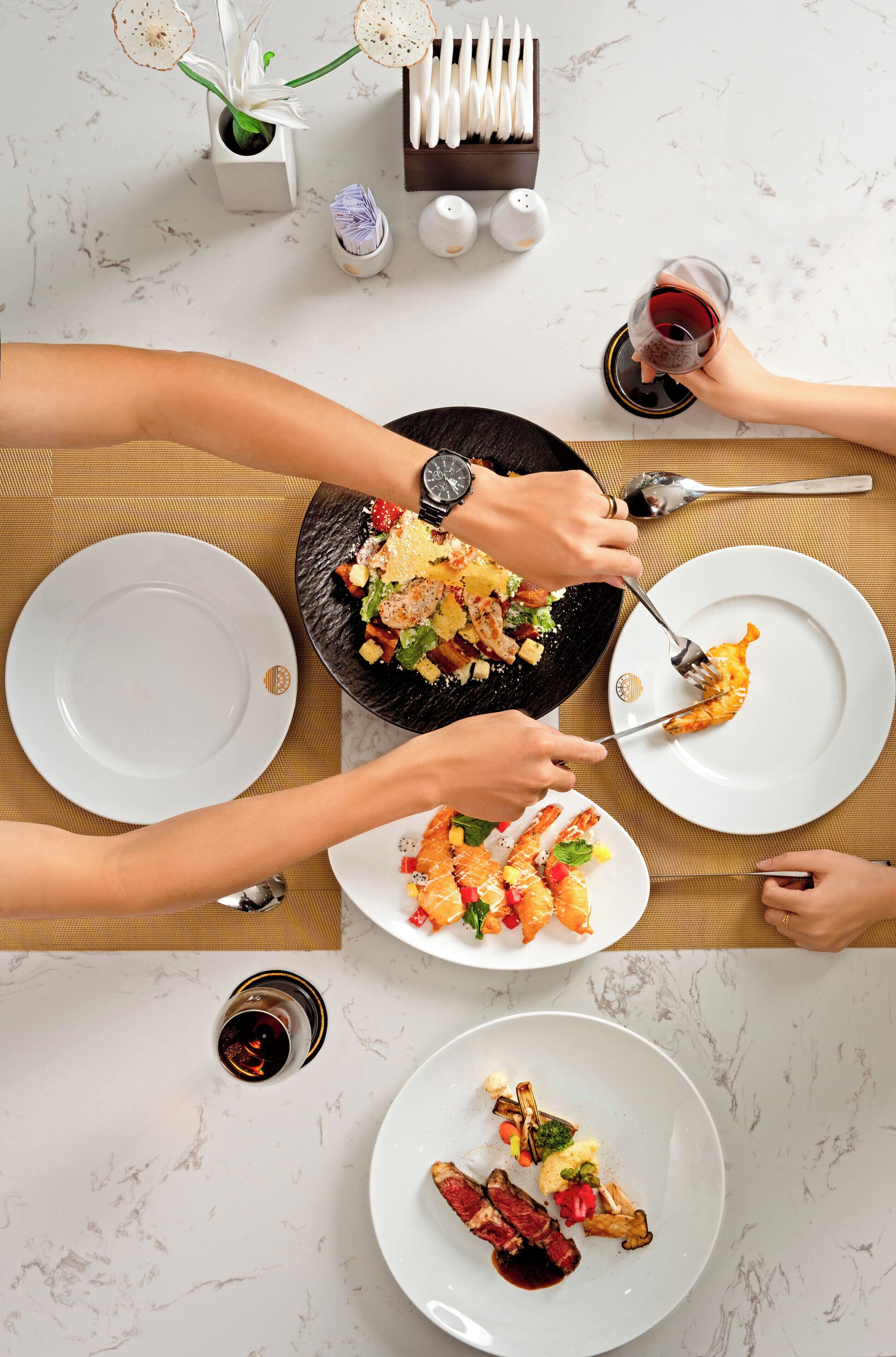 A dining table with multiple plates of food, utensils, and glasses of red wine.
