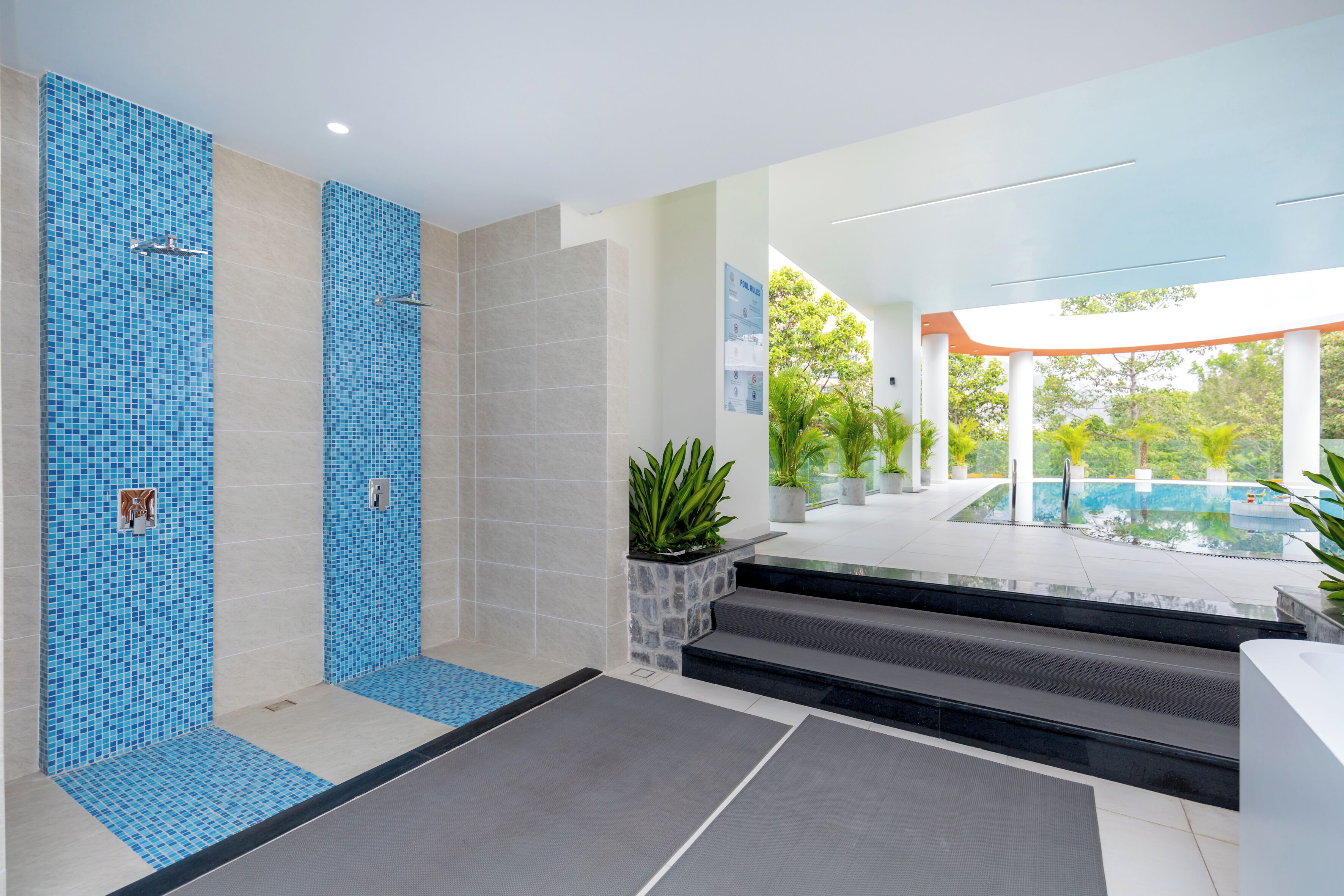 Un baño moderno con ducha de mosaico azul, una maceta de piedra con plantas y vistas a la zona de la piscina.