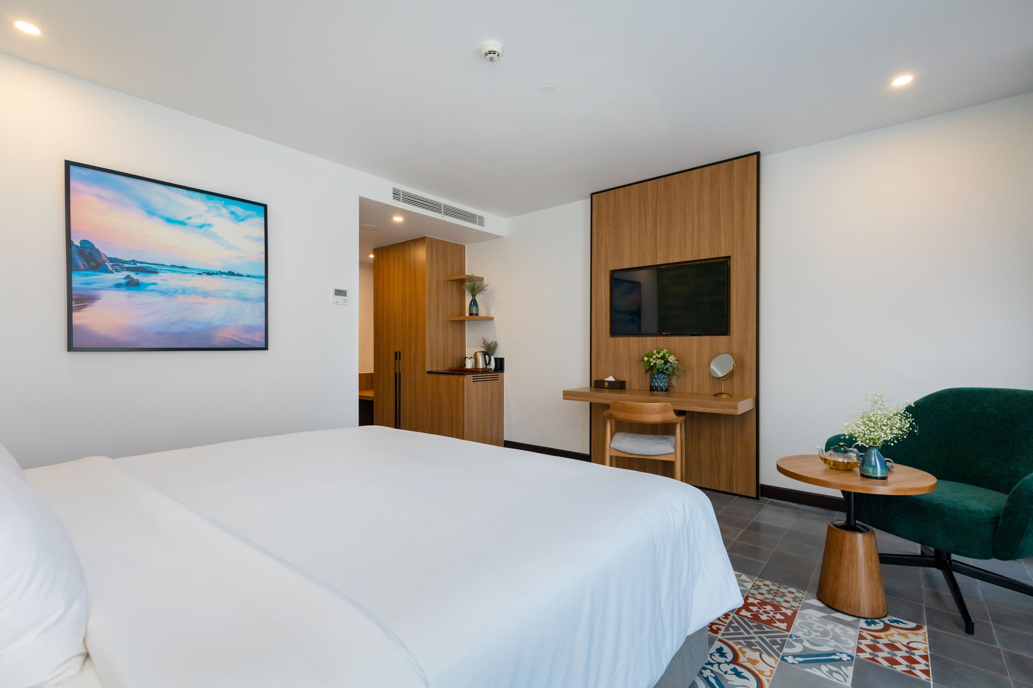 A hotel room with a large bed, a desk with a chair, a small table with a vase, and a painting on the wall.