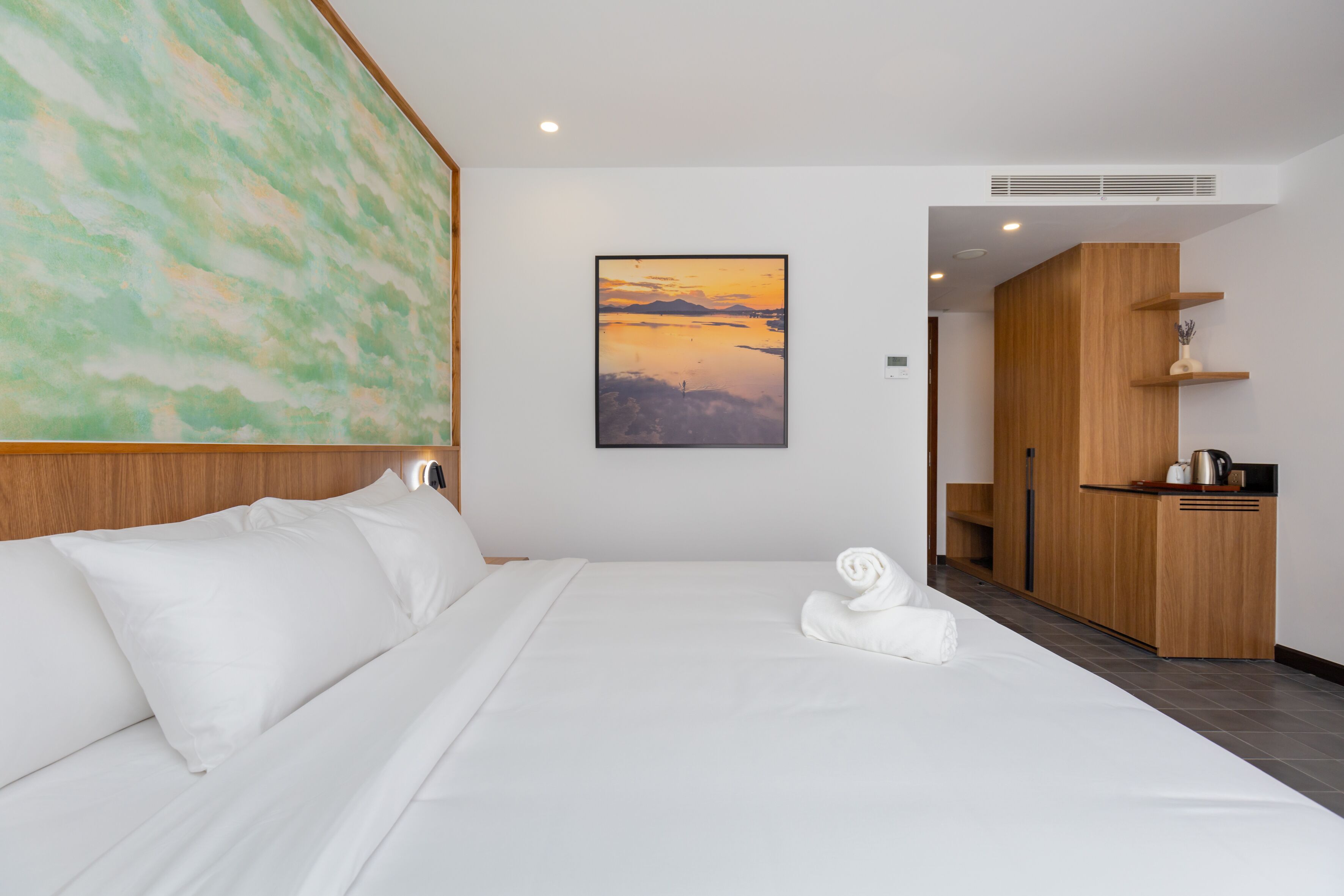 A hotel room with a large bed, a painting of a sunset, a wooden wardrobe, and a small kitchenette.