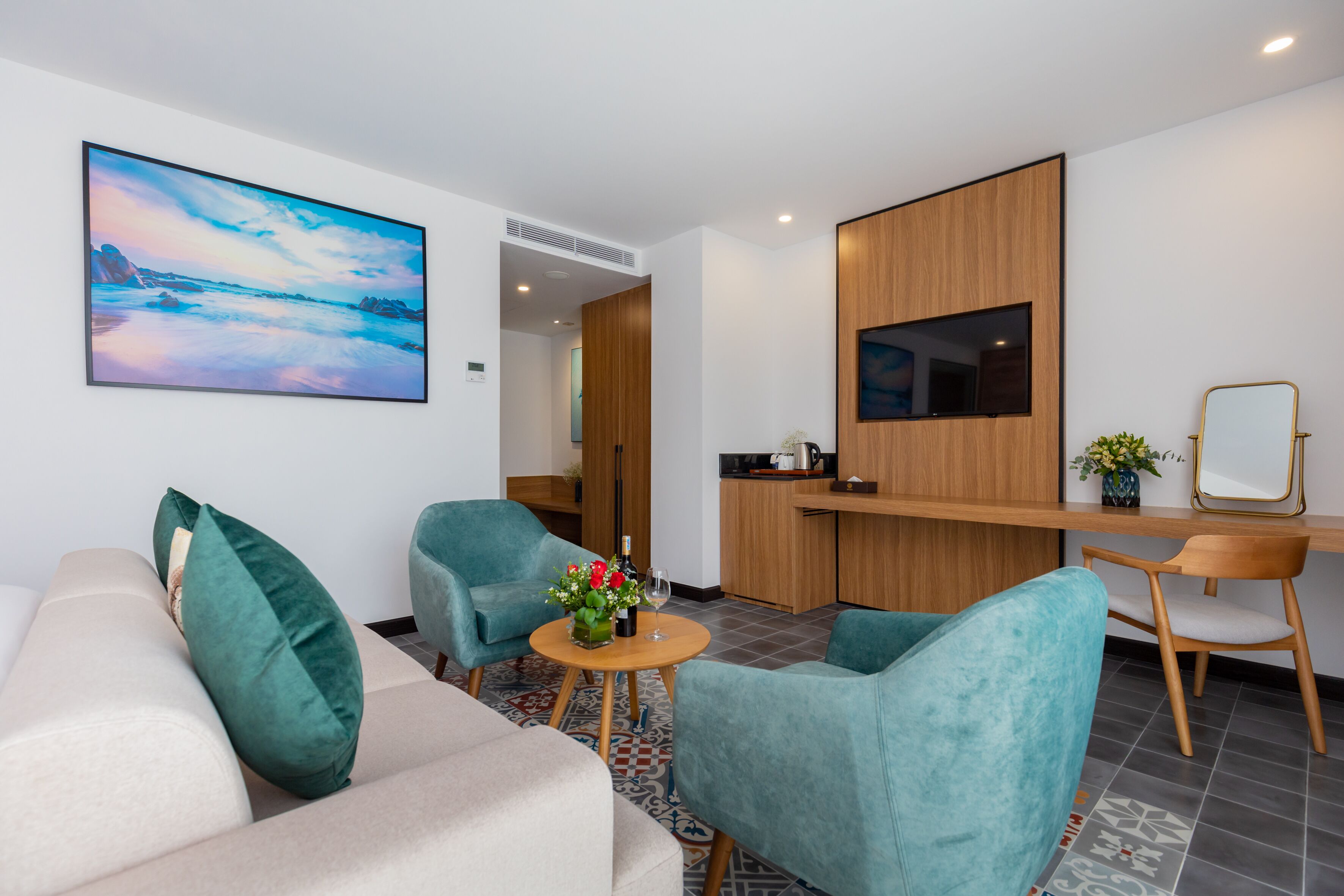 A modern hotel room with a sofa, two armchairs, a small round table with a bottle and flowers, a wooden desk, and a TV mounted on the wall.