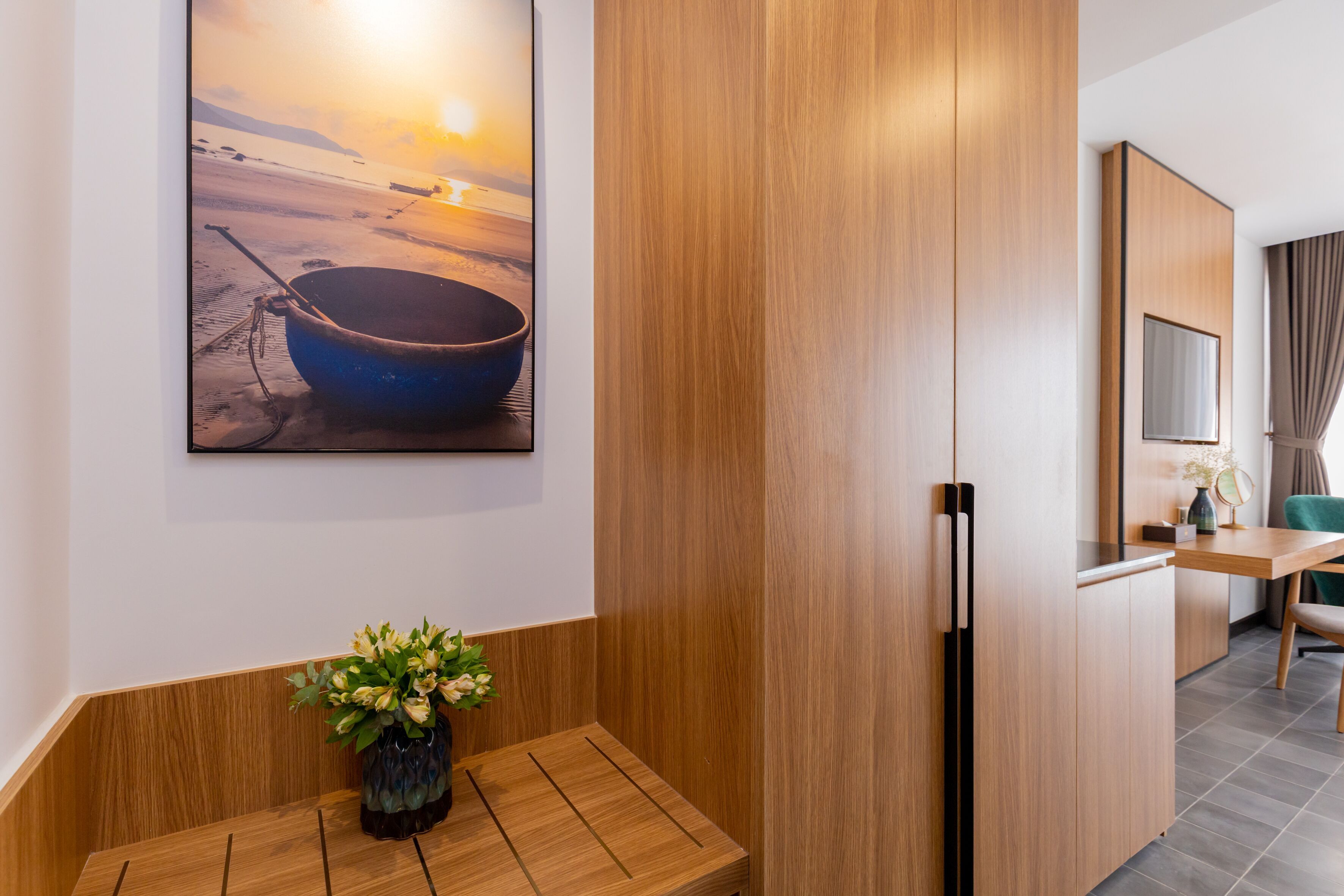 A modern hotel room with a wooden bench, a painting of a boat on a beach, and a wooden cabinet.