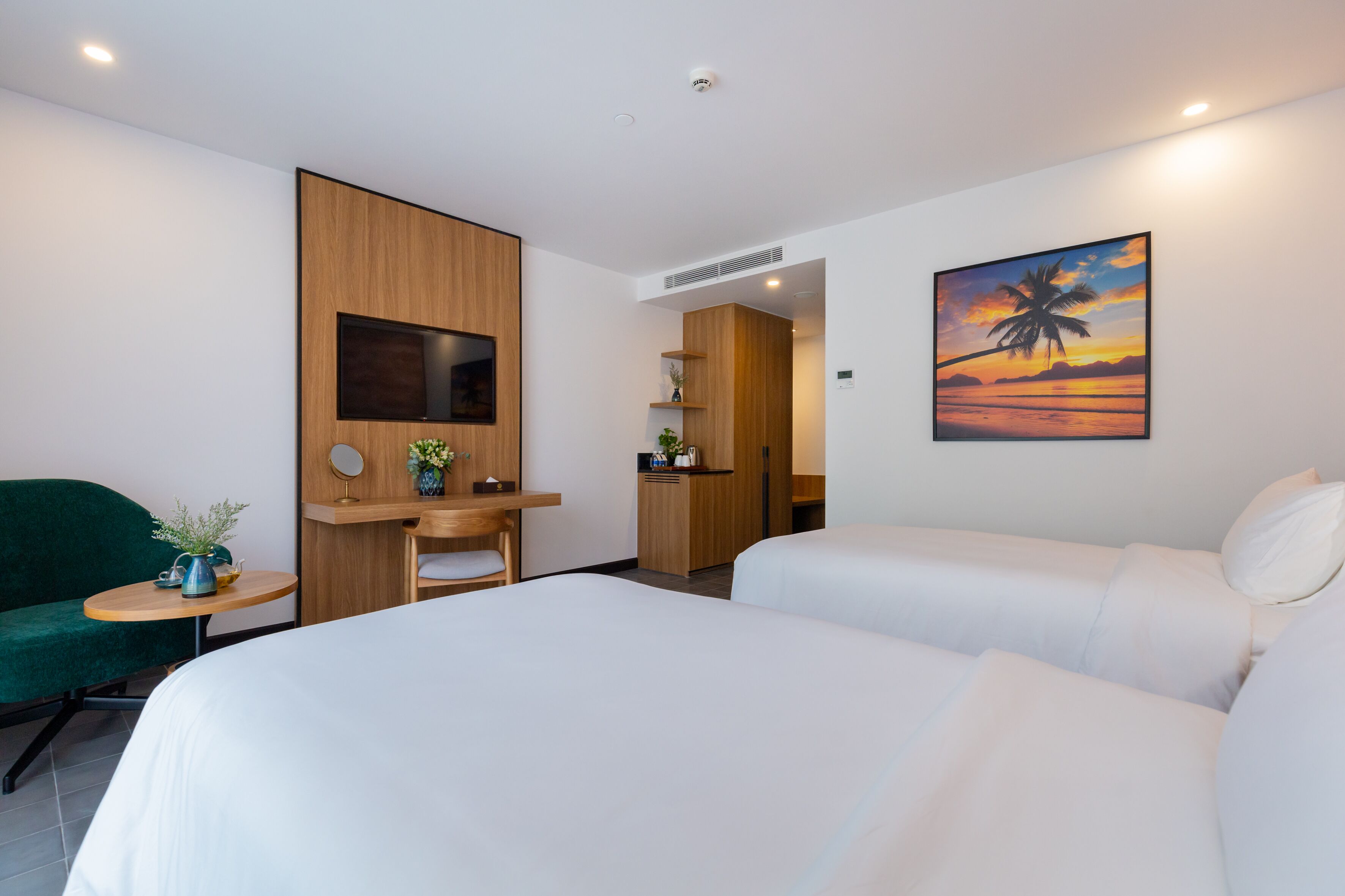 A hotel room with two beds, a green armchair, a small table with a plant, a wooden desk, and a painting of a sunset.
