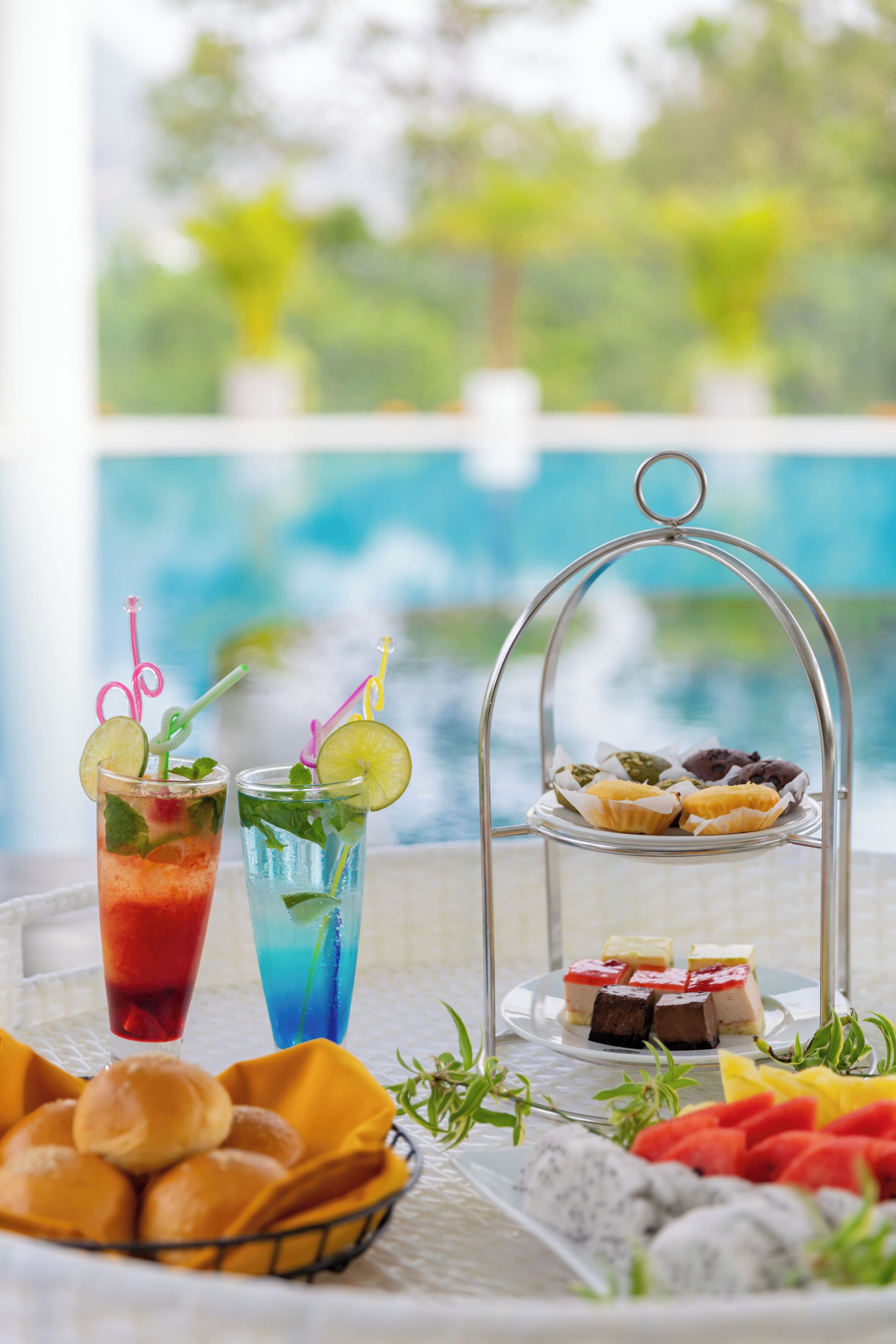 A table with two drinks, a tray of pastries, and a basket of bread rolls.