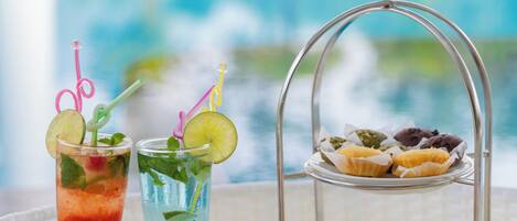 A table with two drinks, a tray of pastries, and a basket of bread rolls.
