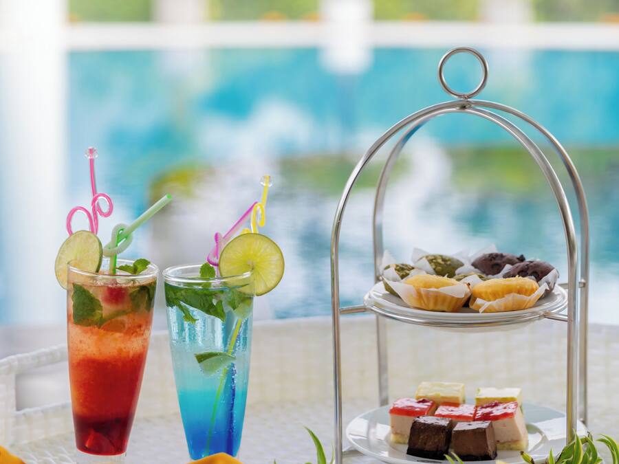 A table with two drinks, a tray of pastries, and a basket of bread rolls.