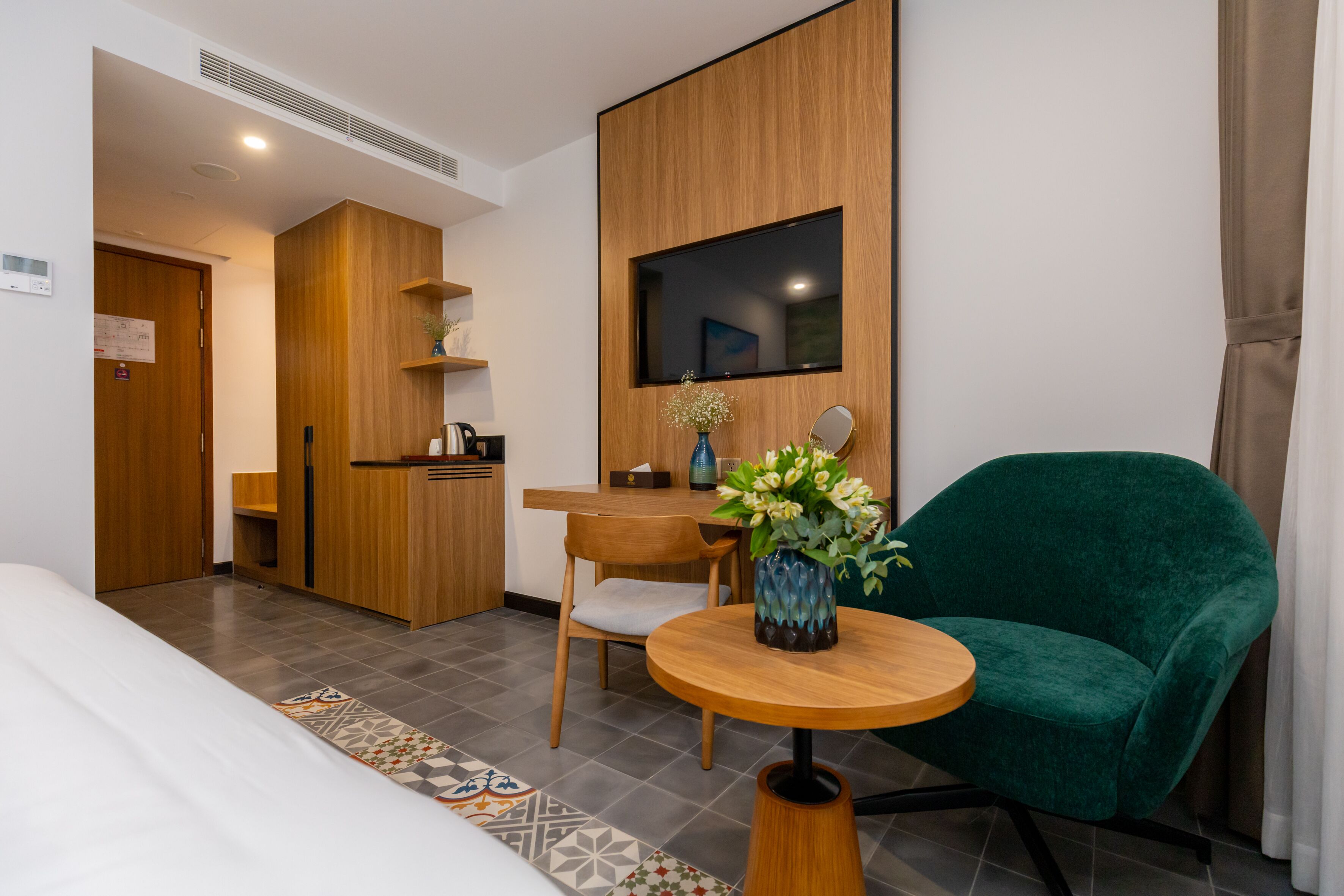 A hotel room with a bed, a green armchair, a wooden table with a vase, a wooden wardrobe, a desk with a chair, and a mounted TV.