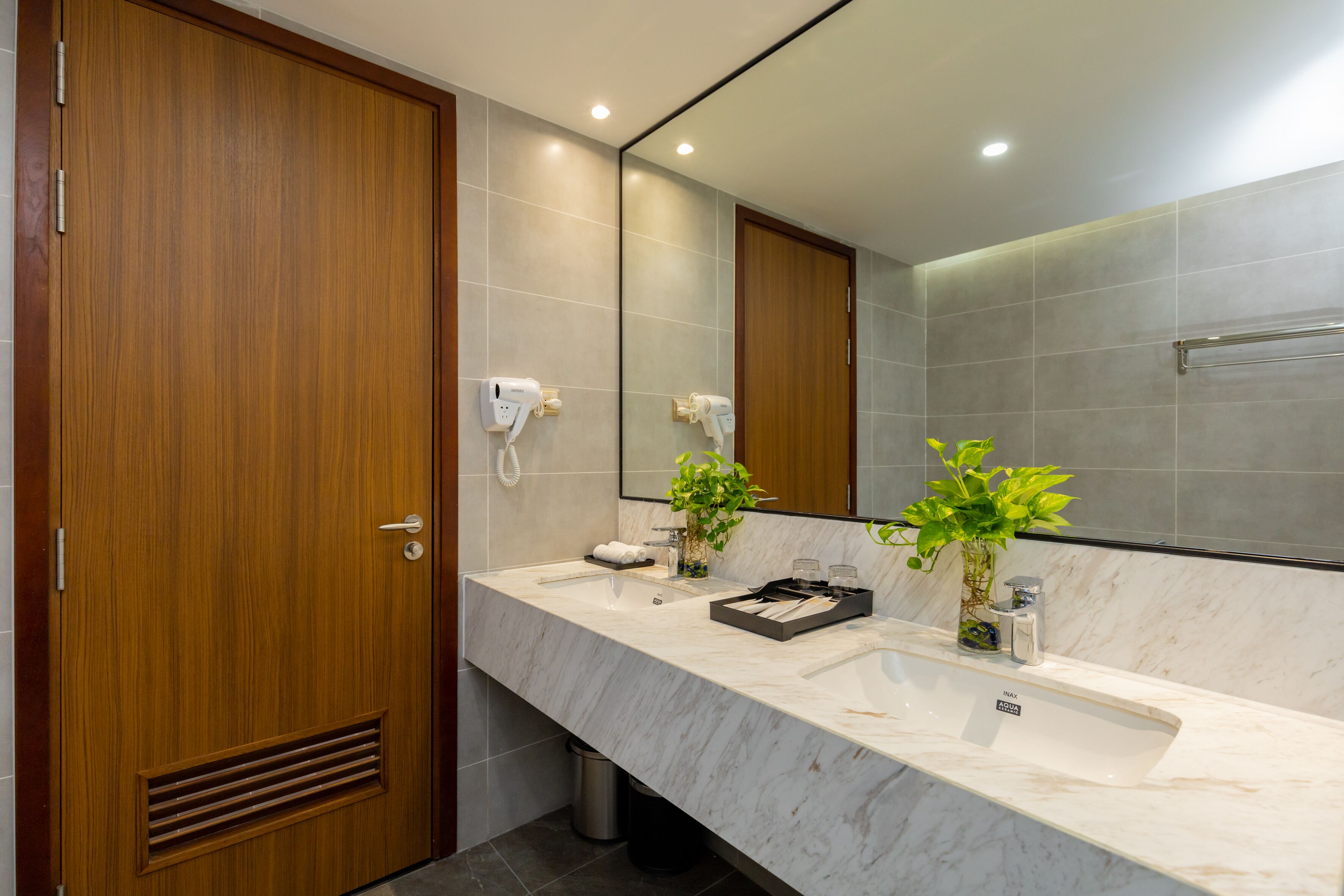 Une salle de bain moderne avec une porte en bois, un lavabo en marbre, deux sèche-cheveux, une plante et un miroir.