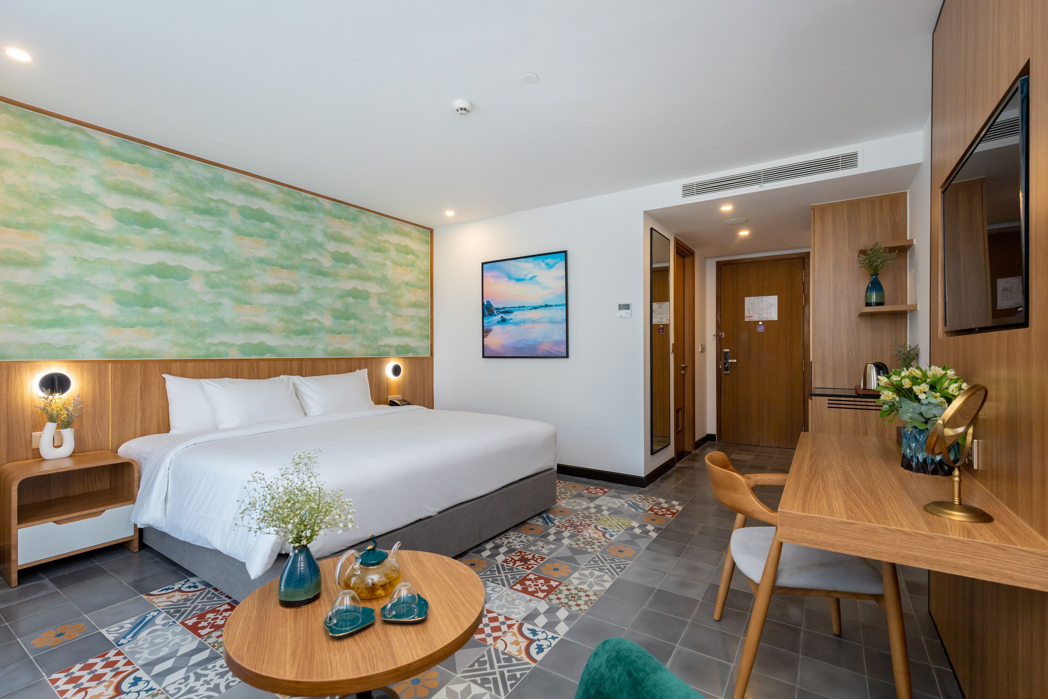 A hotel room with a large bed, a desk with a chair, a round table with a vase, and a painting on the wall.