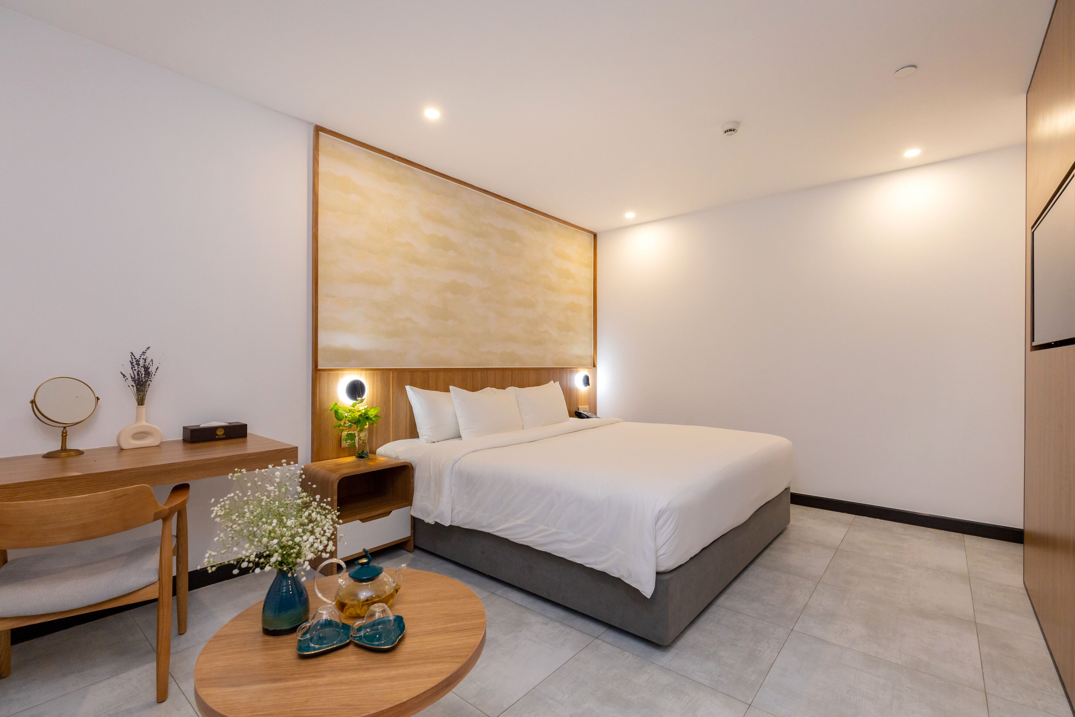 A modern hotel room with a large bed, a wooden desk, a round coffee table with decorative items, and a wall-mounted artwork.
