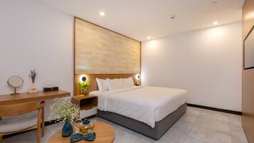 A modern hotel room with a large bed, a wooden desk, a round coffee table with decorative items, and a wall-mounted artwork.