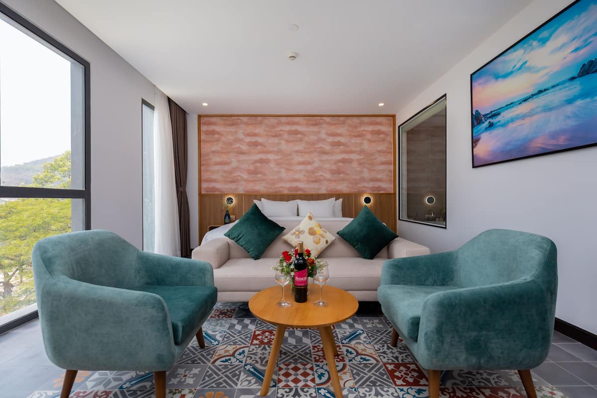 A modern hotel room with a large bed, two teal armchairs, a small round table with a bottle of wine and glasses, and a large window offering a view of trees.