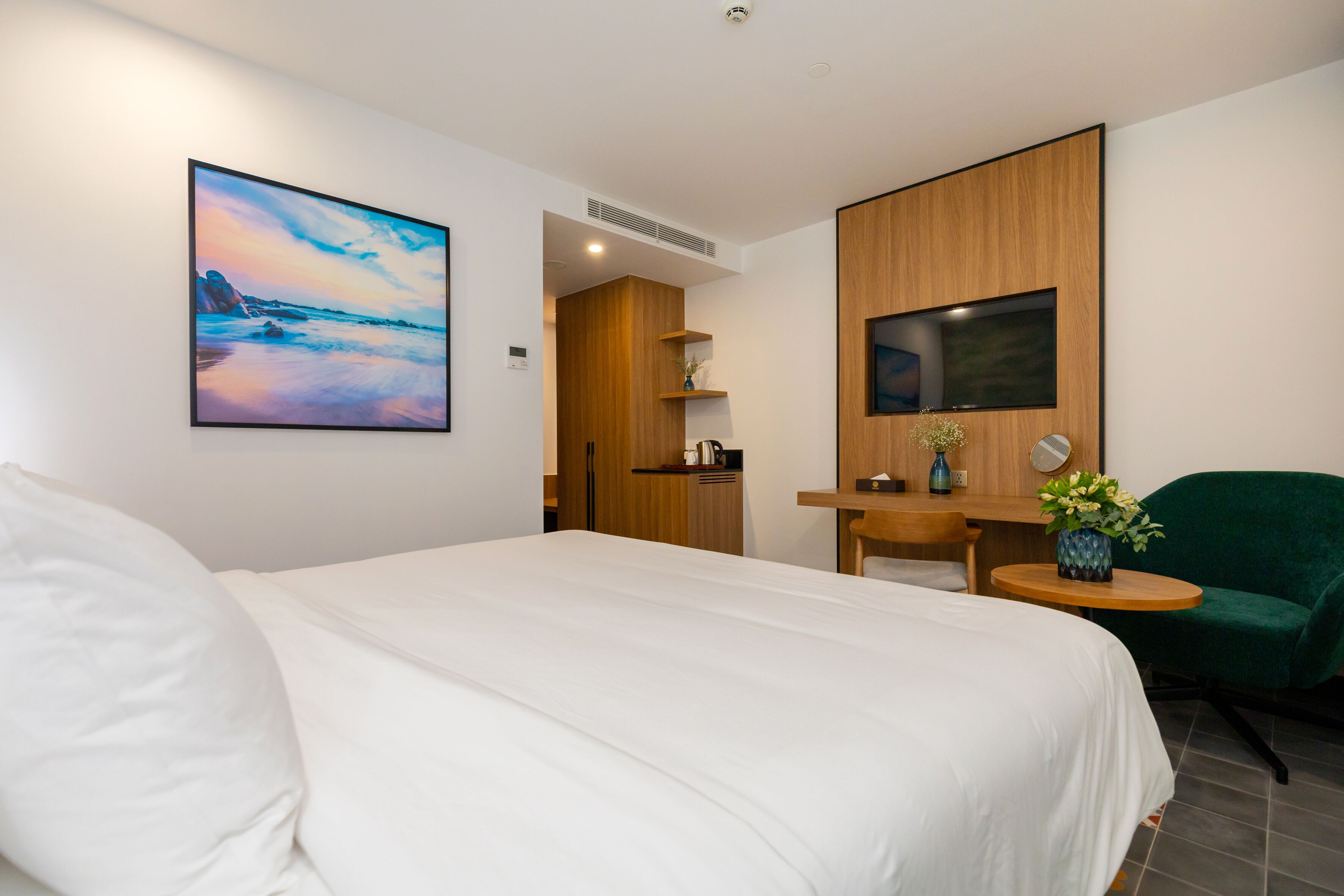 A hotel room with a bed, a desk, a chair, a painting, and a small table with a vase.