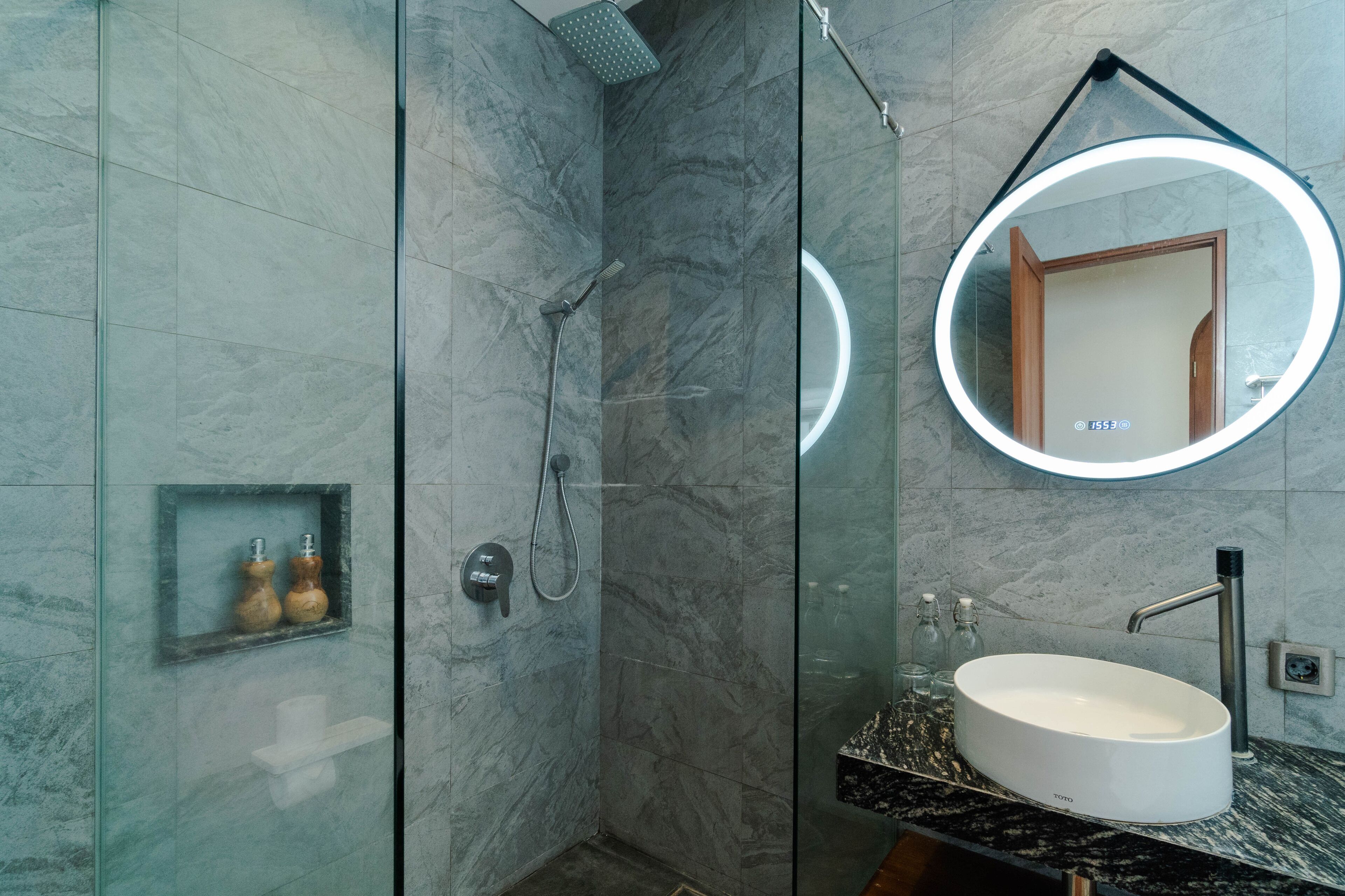 A modern bathroom with a glass-enclosed shower, a round mirror with a light, and a marble-tiled sink.