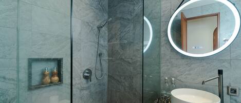A modern bathroom with a glass-enclosed shower, a round mirror with a light, and a marble-tiled sink.