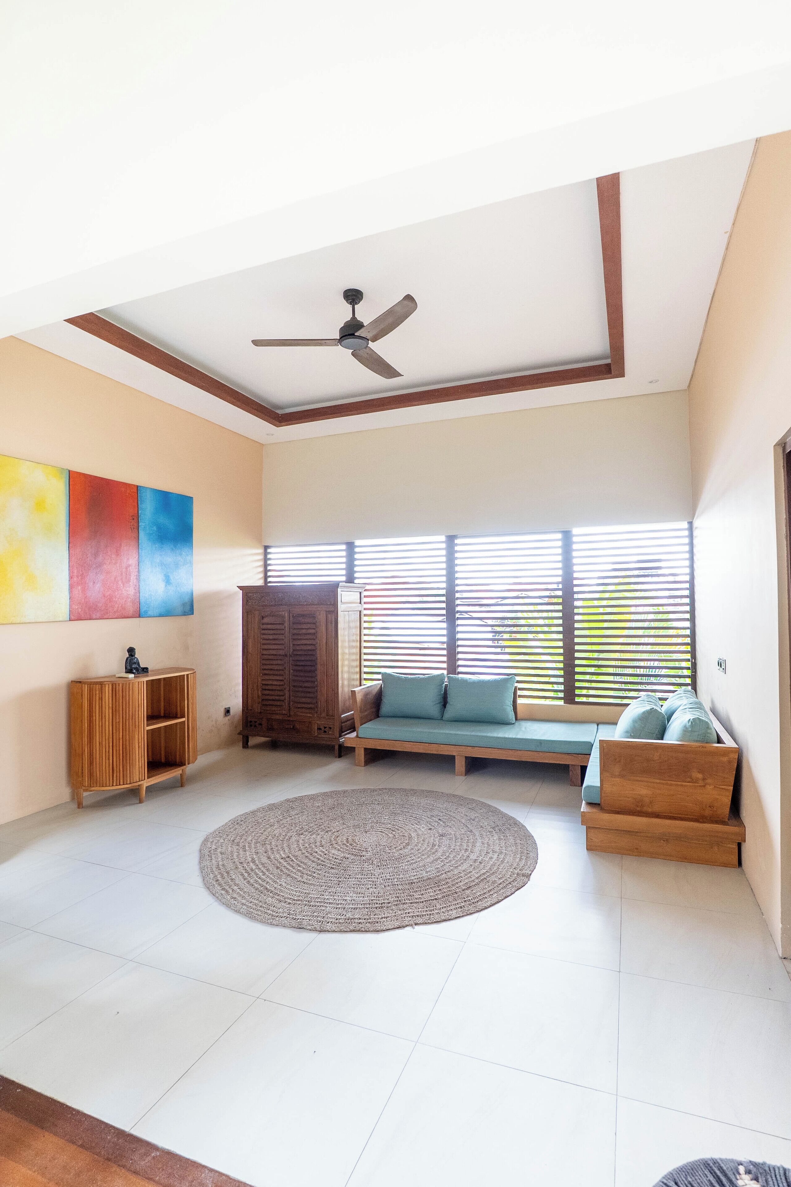 Un amplio salón con ventilador de techo, un sofá, un mueble de madera y un gran ventanal con persianas.