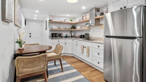 Een moderne keuken met witte kasten, een roestvrijstalen koelkast en een eethoek met rieten stoelen en een gestreept vloerkleed.