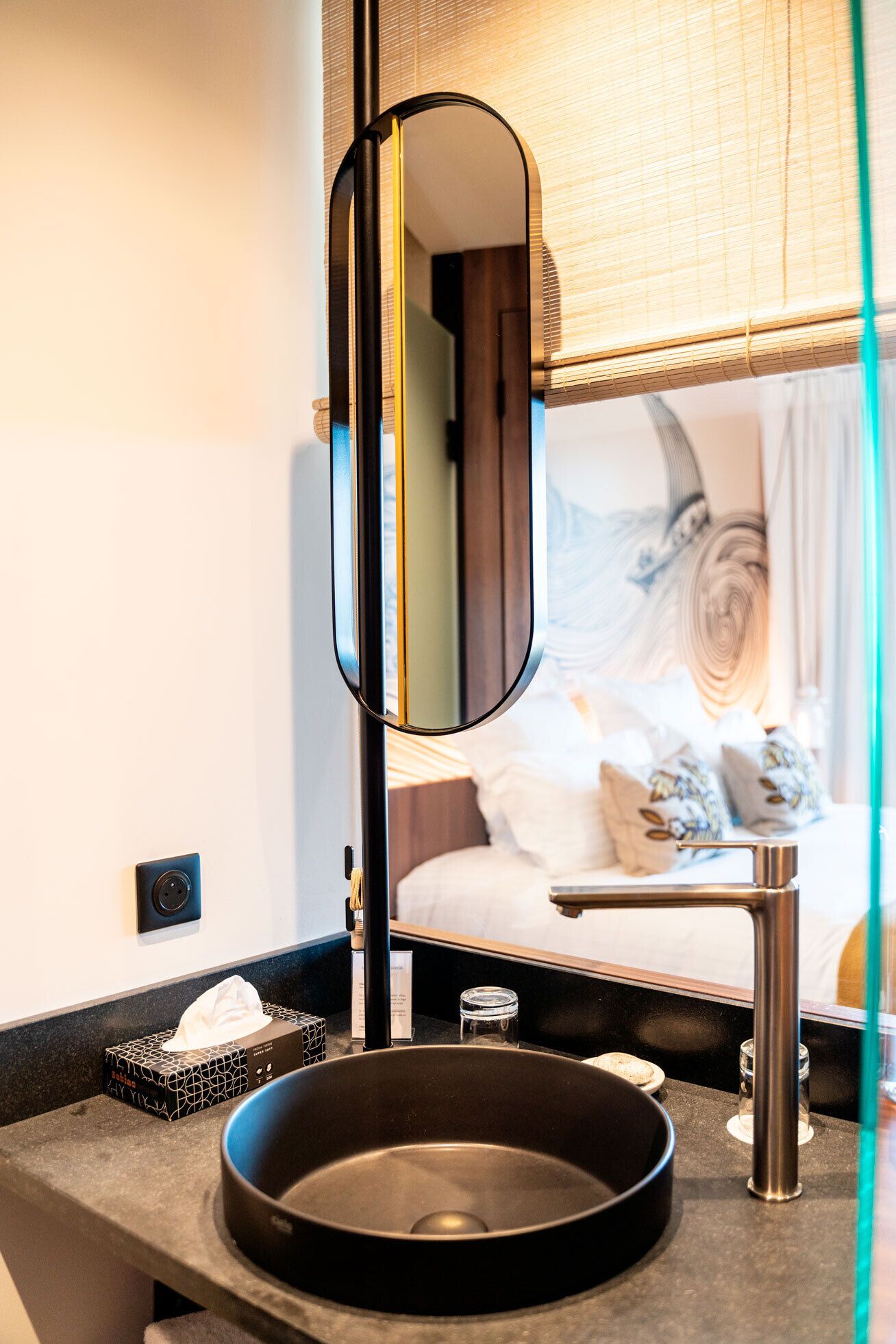 A bathroom with a round sink, a mirror, a towel dispenser, and a bed visible in the reflection.