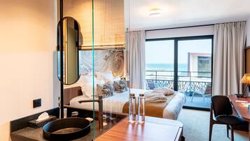Habitación de hotel con cama, escritorio, silla, baño con lavamanos y un amplio ventanal con vista a la playa y al océano.