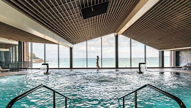 An indoor swimming pool with a view of the sea, large windows, and a modern ceiling design.