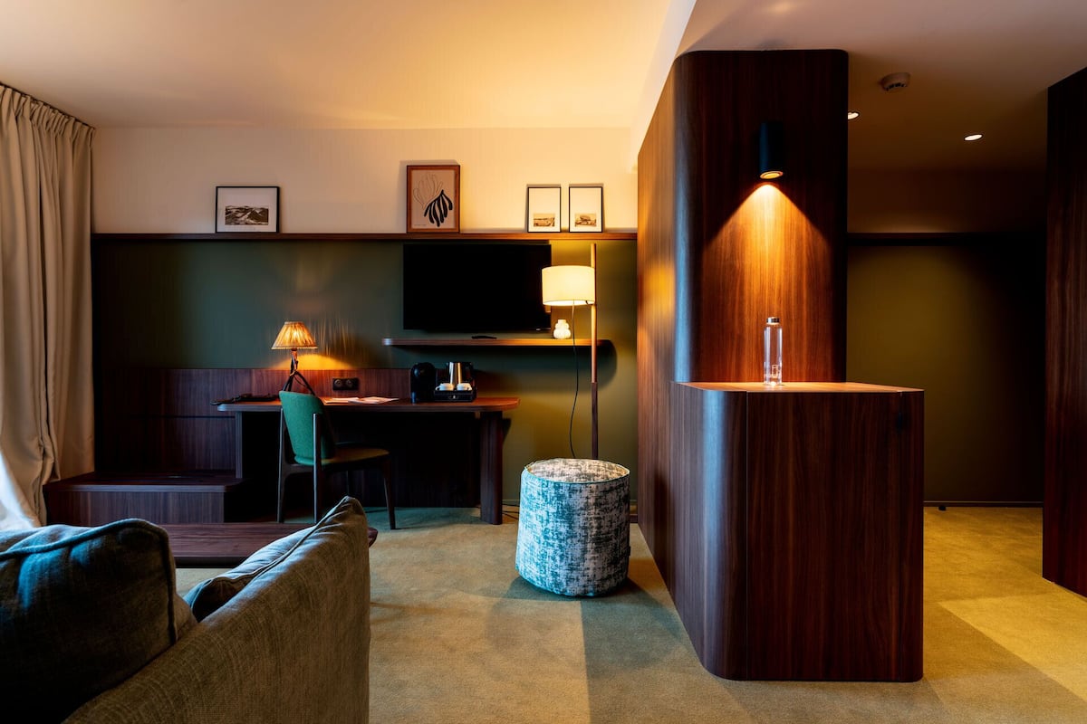 A modern hotel room with a wooden desk, a television, a lamp, and a small round ottoman.