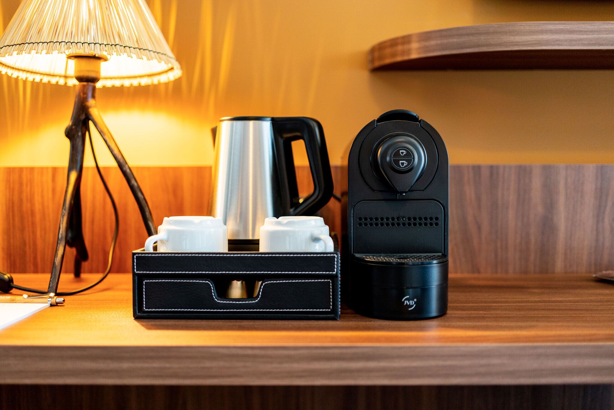 Une machine à café, une bouilloire et un porte-tasse sur une table en bois, avec une lampe en arrière-plan.