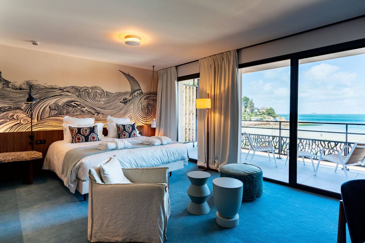 A hotel room with a large bed, a view of the sea, and a balcony with chairs.