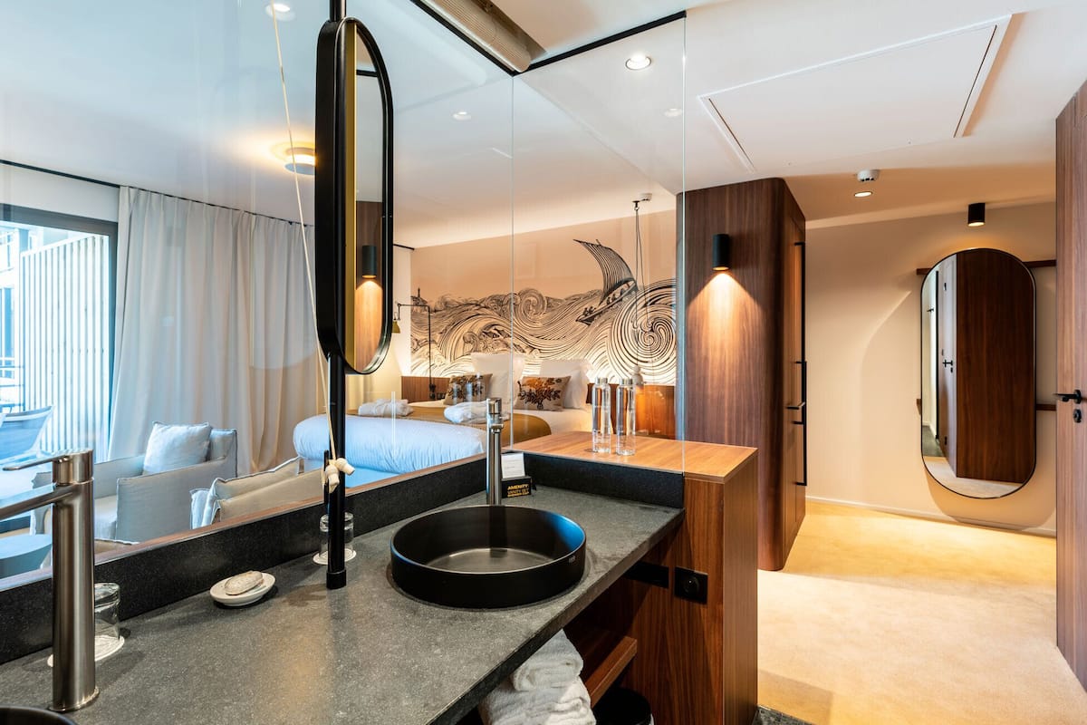 A modern hotel room with a large bed, a round sink, a mirror, and a view of the city.