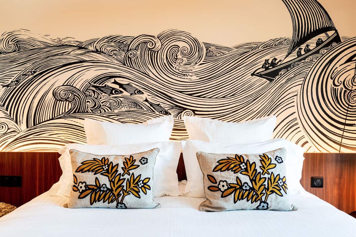 A bed with white linens and two decorative pillows with floral embroidery, set against a wall with a large black and white wave and boat mural.