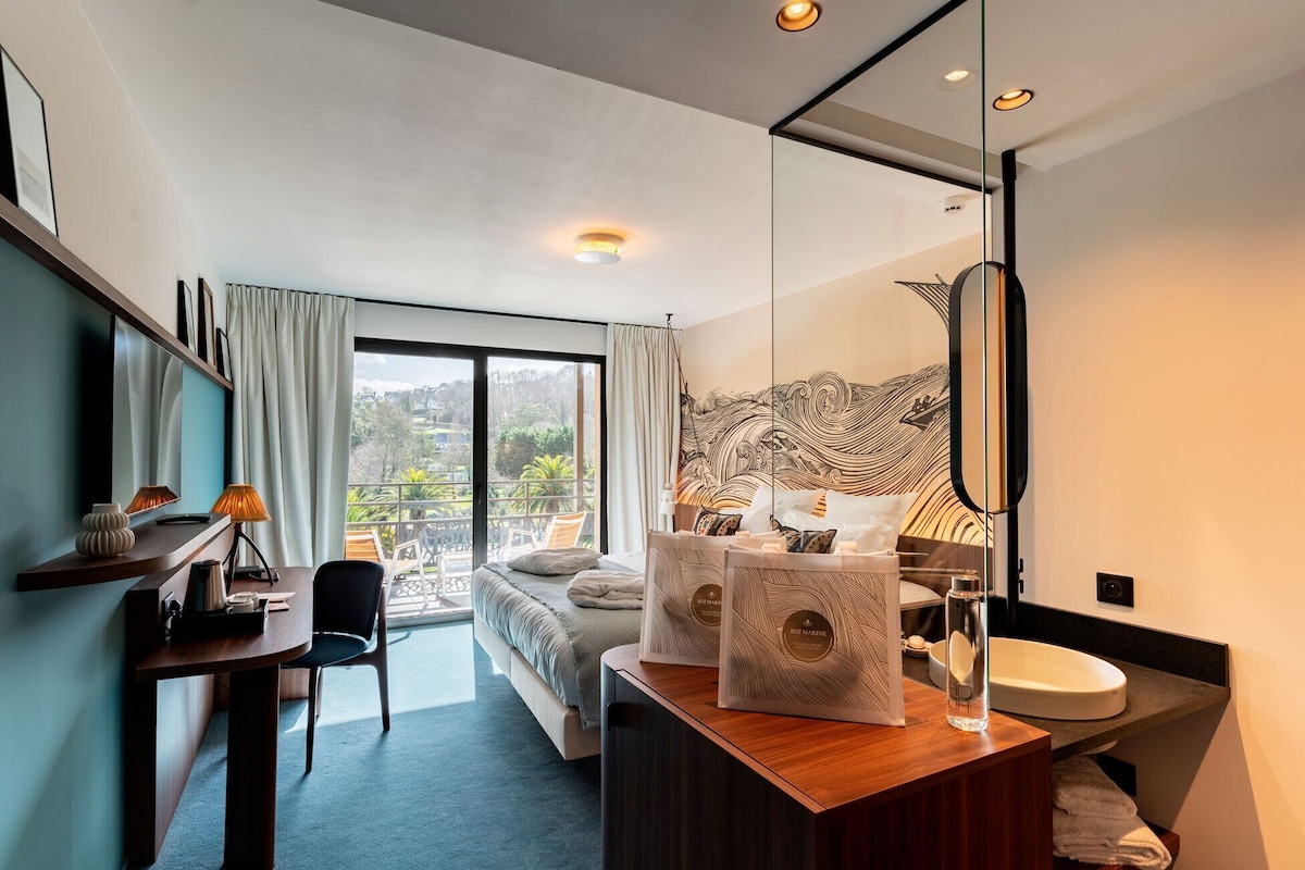A modern hotel room with a bed, a desk, a chair, a bathroom, and a view of the outdoors.