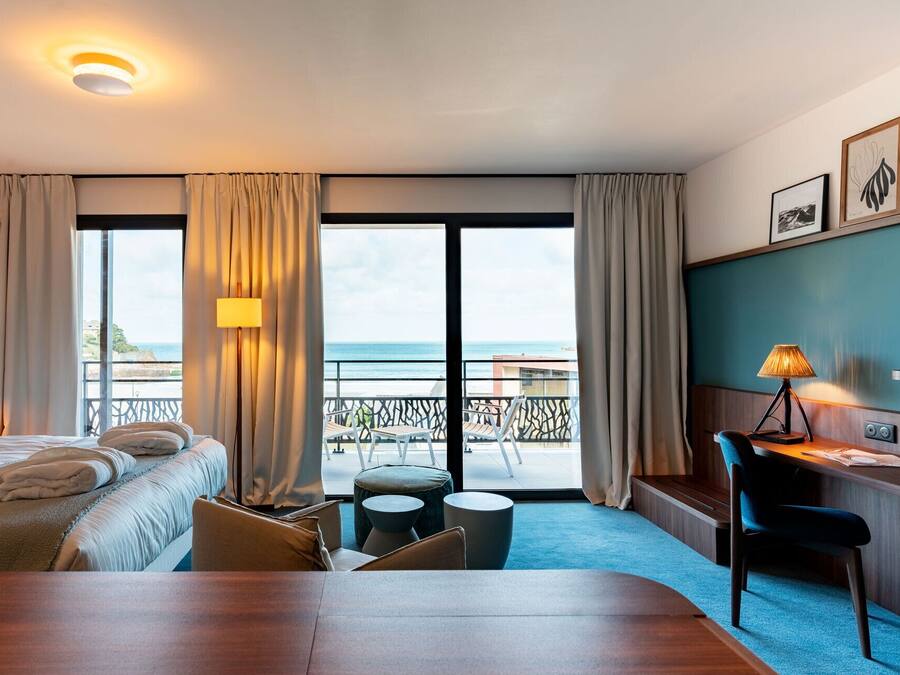 A hotel room with a bed, a desk, a chair, a small table, and a view of the sea.