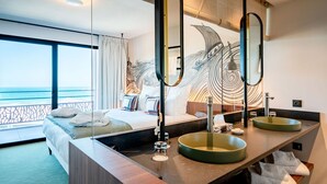 A hotel room with a large bed, two round mirrors, a vanity with a green sink, and a view of the sea through large windows.