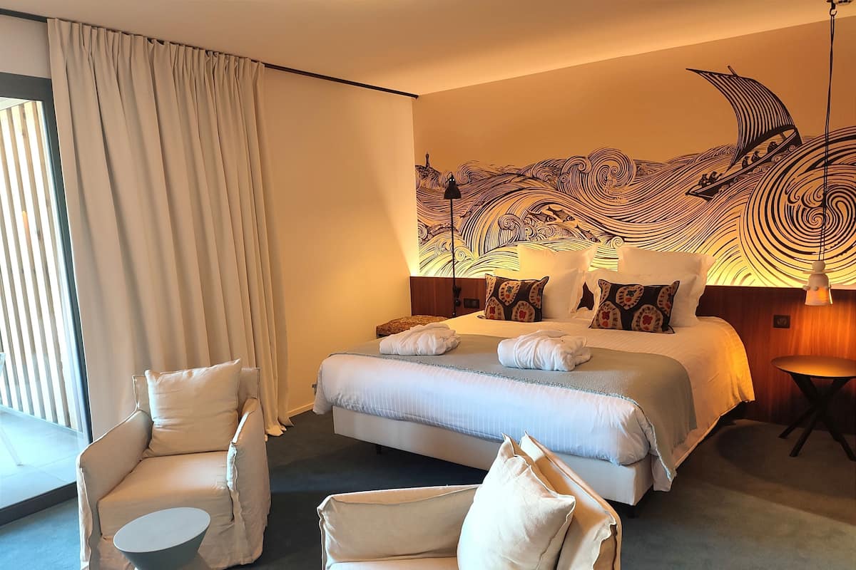 A hotel room with a bed, two chairs, a small table, a large window with curtains, and a wall mural.