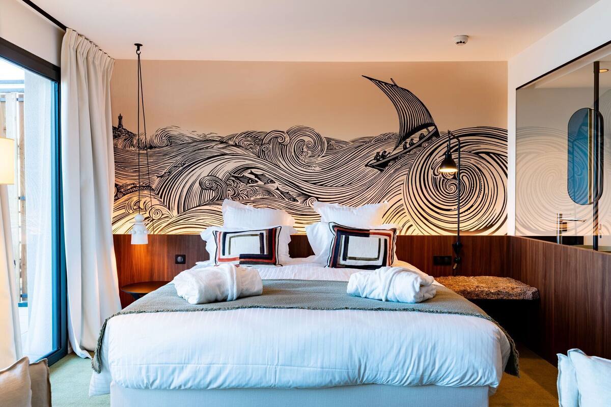 A hotel room with a large bed, a wall mural, a mirror, and a window with curtains.