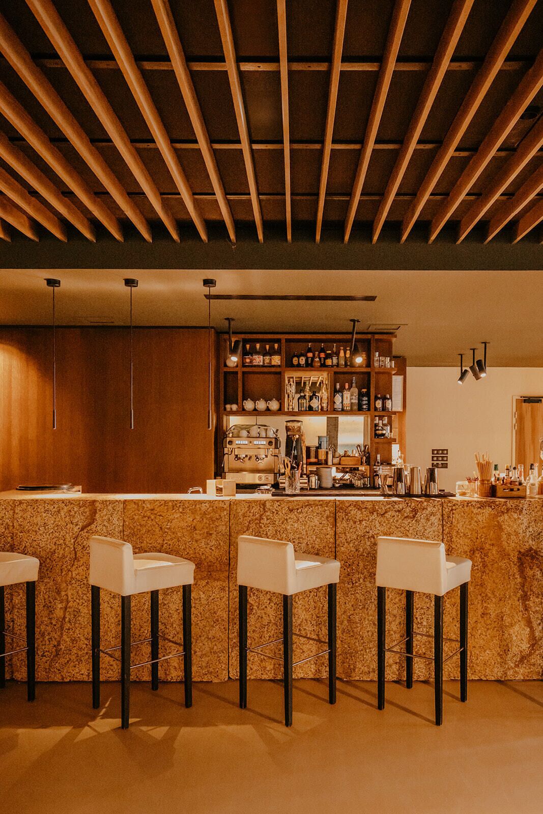 Un espace bar avec un plafond en bois, des tabourets de bar et un comptoir où sont disposées diverses bouteilles et une machine à café.