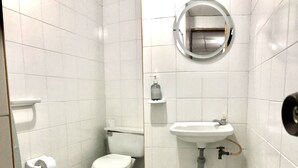 Une petite salle de bain aux murs recouverts de carreaux blancs, équipée de toilettes, d’un lavabo, d’une poubelle et d’un miroir.