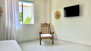 Une pièce comprenant un lit, une chaise en bois, un téléviseur fixé au mur et une fenêtre avec des rideaux.