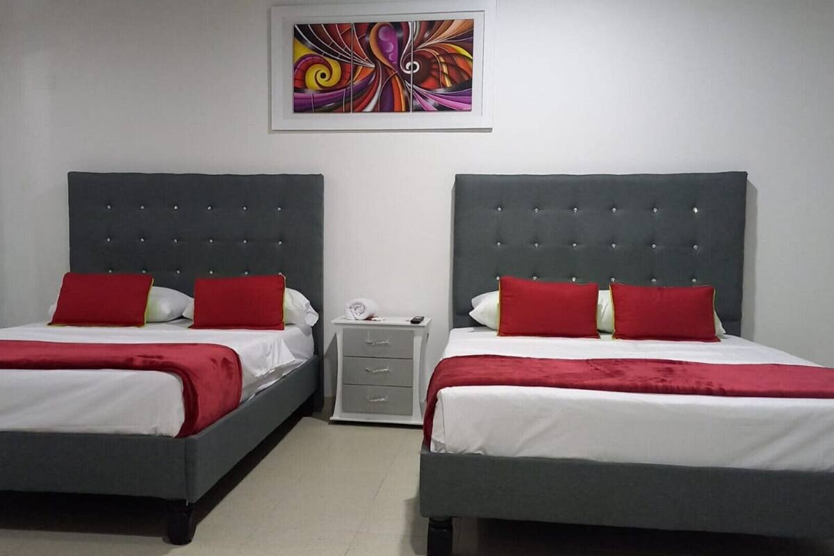 Habitación de hotel con dos camas, cada una con cabecero gris y ropa de cama roja. Hay una mesita de noche pequeña con una lámpara y un cajón. Un cuadro abstracto y colorido cuelga en la pared sobre las camas.