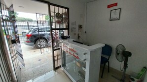 Un mostrador de recepción con separador de vidrio, un coche estacionado afuera y un ventilador a la derecha.
