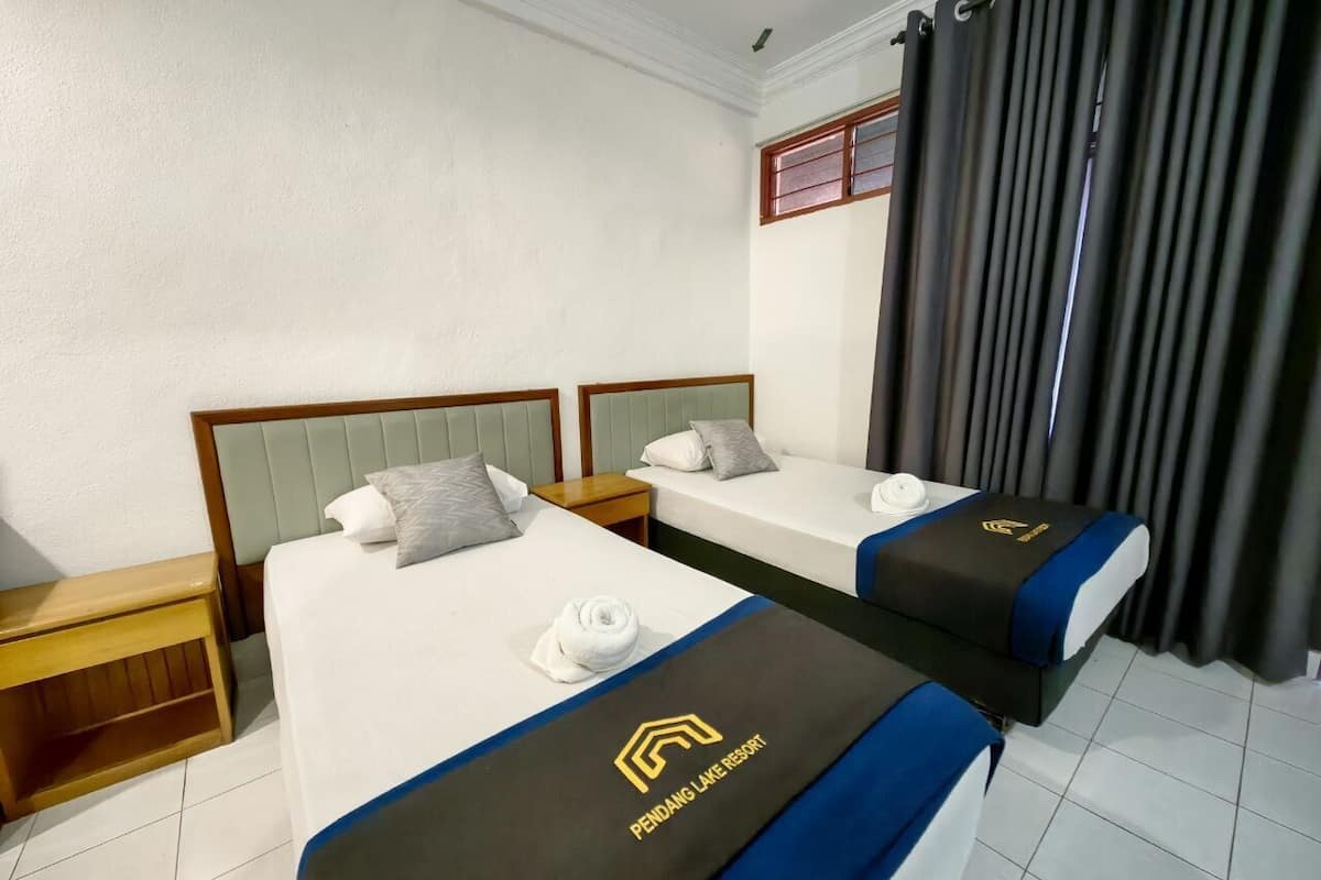 A hotel room with two beds, each with a pillow and a rolled towel. The beds have a logo on the bedspread. There is a nightstand and a window with curtains.