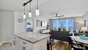 Une cuisine moderne avec un comptoir en marbre, des armoires blanches et des luminaires suspendus. On y trouve un coin repas avec une table et des chaises, ainsi qu’un salon avec un canapé gris et une vue sur l’océan.