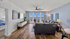 Un salon moderne avec un canapé gris, une table à manger et un ventilateur de plafond. Il y a un balcon avec vue sur l’océan.