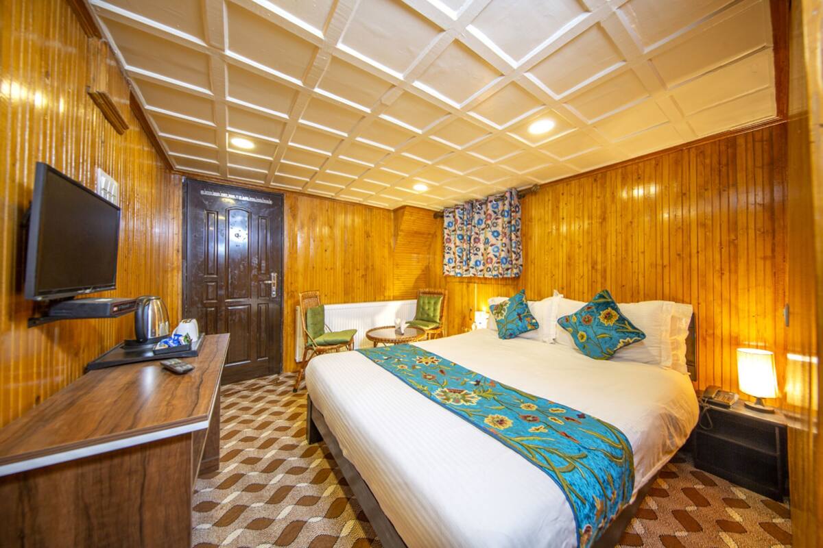 Quarto de hotel com cama, escrivaninha com televisão, cadeira, mesinha e chão estampado.