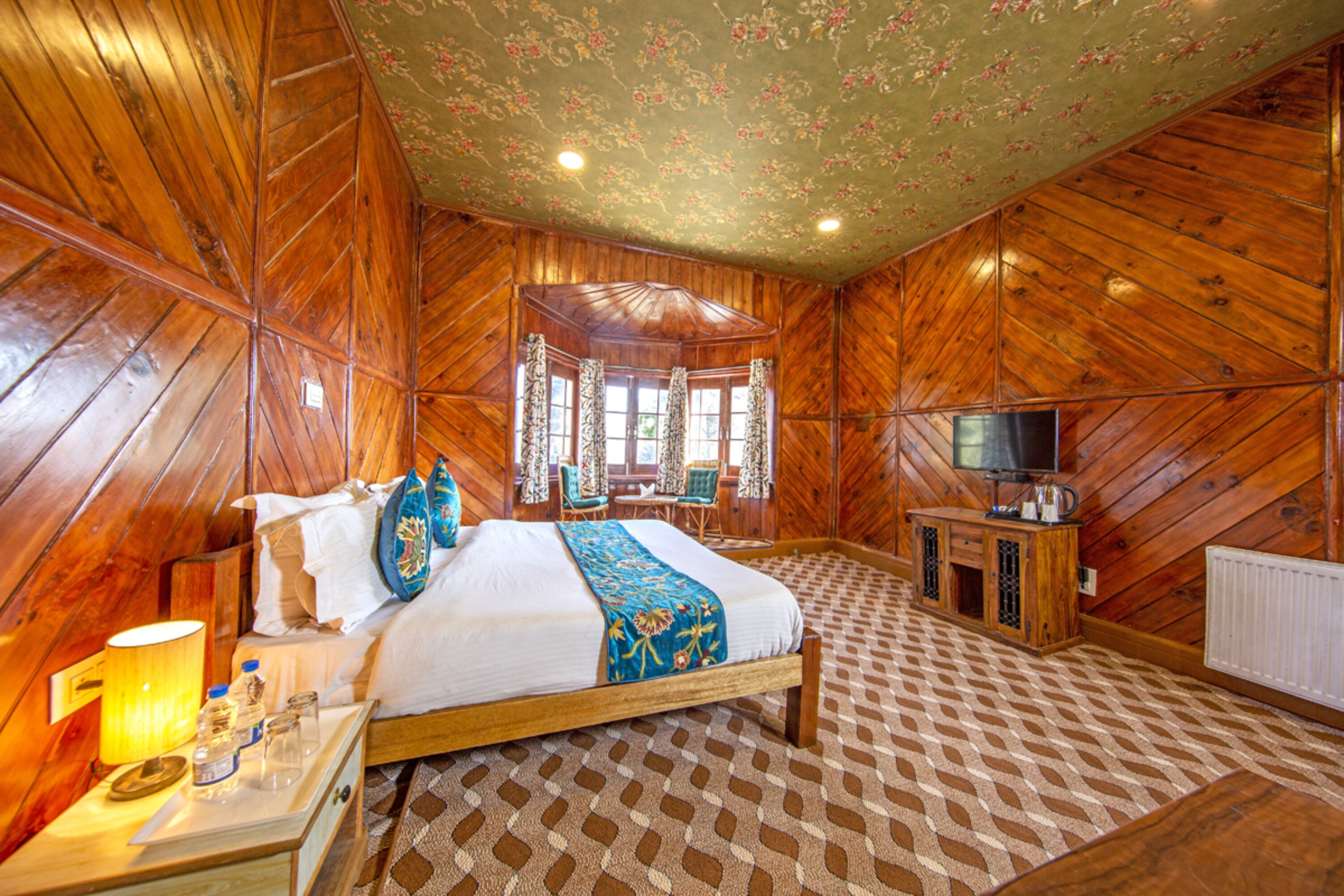 A wooden-paneled room with a bed, a television, and a table with a lamp and glasses.