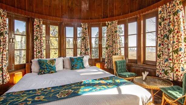 A wooden cabin bedroom with a large bed, floral curtains, and a view of trees. - Gulmarg Ski Hill Resort (Baramula)