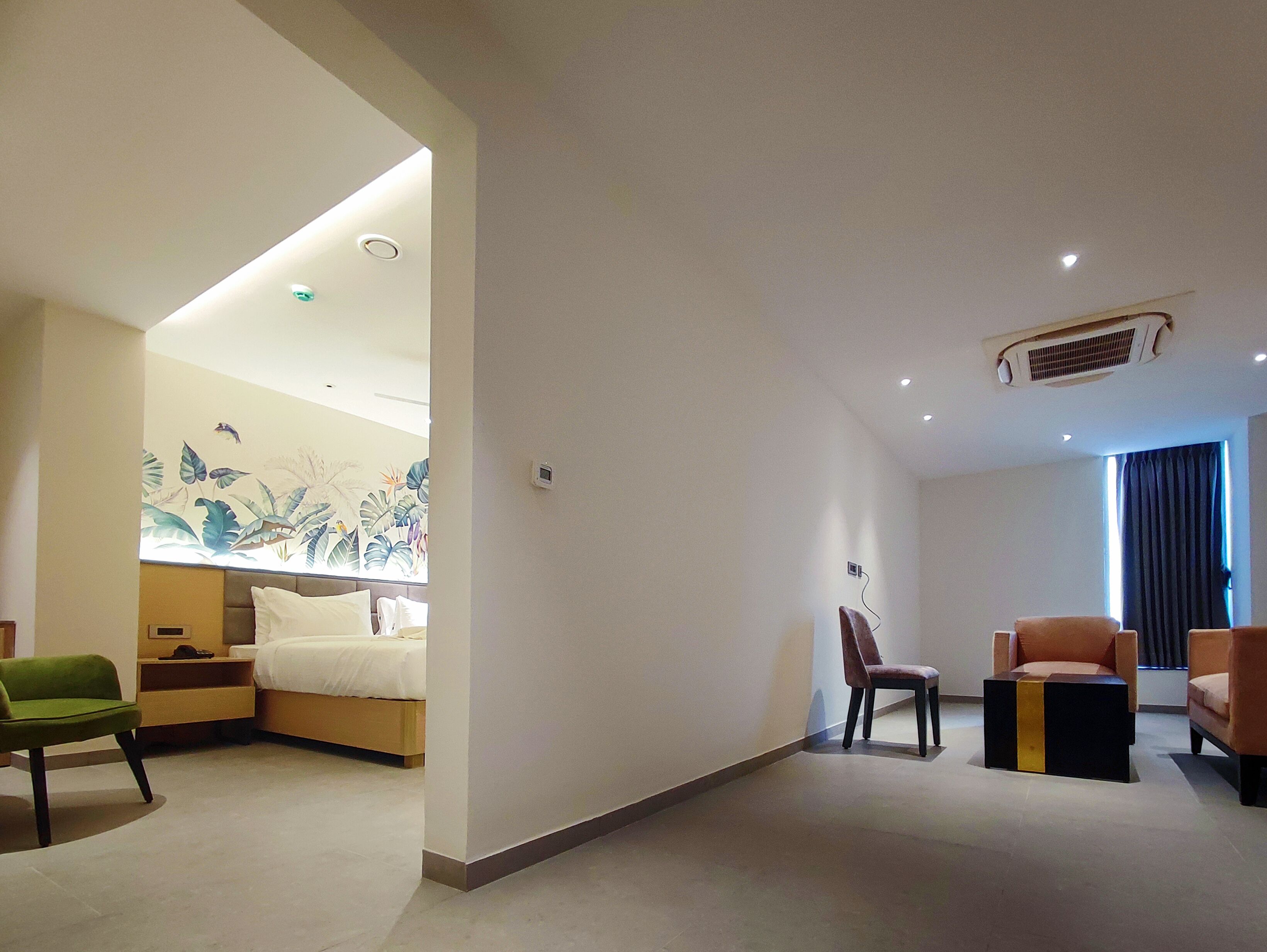 A modern hotel room with a bed, a green chair, a brown armchair, a black and gold side table, and a wall mural.