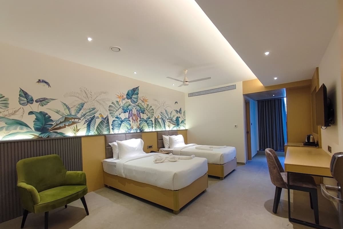 Ein Hotelzimmer mit zwei Betten, einem grünen Sessel, einem Schreibtisch mit Stuhl, einem Deckenventilator und einem Wandbild.